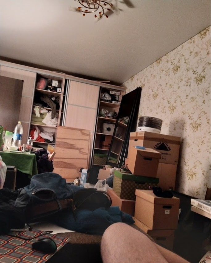 📷 Захламлённая комната при переезде или уборке: стопки картонных коробок, открытый шкаф-купе с вещами, полки, цветочные обои, полосатый ковёр. На переднем плане ноги лежащего человека.