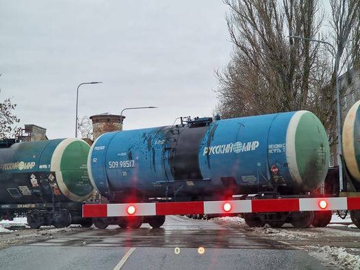 📷 Железнодорожные цистерны голубого цвета с надписью «РУССКИЙ МИР» на путях у переезда со шлагбаумом, серый день.
