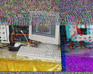 Сильно зашумлённое SSTV-изображение: едва различим компьютерный стол с монитором и клавиатурой; справа — другой объект, возможно радиооборудование; сильный цветной шум