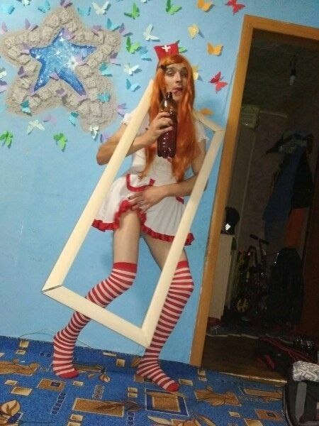 📷 Тот же косплей медсестры: молодой мужчина в белом платье, красно-белых полосатых чулках, рыжем парике; стоит внутри белой прямоугольной рамки; синие стены, стеллаж на фоне.