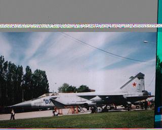 SSTV-изображение с небольшим шумом: военный реактивный самолёт (предположительно МиГ-29, бортовой номер «07», красная звезда на хвосте) стоит на аэродроме; деревья и голубое небо на заднем плане