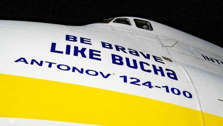 📷 Нос самолёта с надписью крупными синими буквами «BE BRAVE LIKE BUCHA ANTONOV 124-100» на белом корпусе с жёлто-синей полосой (цвета украинского флага), ночная съёмка.