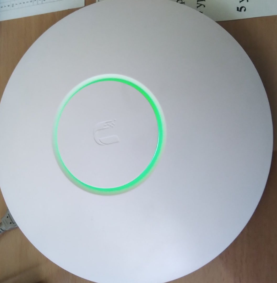 📷 Круглая белая точка доступа Ubiquiti UniFi с зелёным световым кольцом и логотипом Ubiquiti в центре, лежит на деревянной поверхности