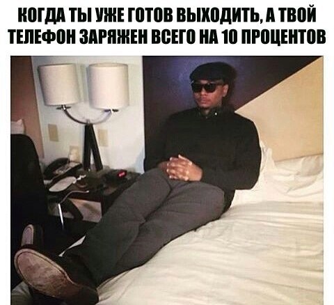 Мем: белая подпись сверху «КОГДА ТЫ УЖЕ ГОТОВ ВЫХОДИТЬ, А ТВОЙ ТЕЛЕФОН ЗАРЯЖЕН ВСЕГО НА 10 ПРОЦЕНТОВ»; фото — темнокожий мужчина в тёмном пальто и кепке, в солнцезащитных очках, сидит откинувшись на белое покрывало кровати в гостиничном номере; за ним лампа и тумбочка